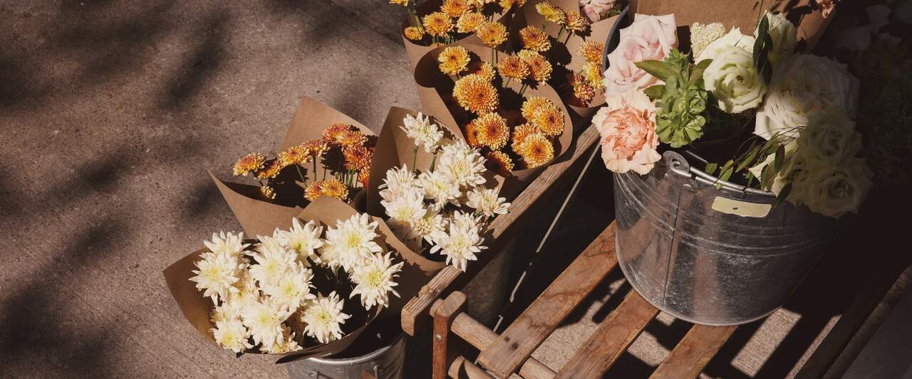Kraft-wrapped bunches of white and orange chrysanthemums and pale roses in metal buckets on a wooden bench, leaf shadows across the pavement