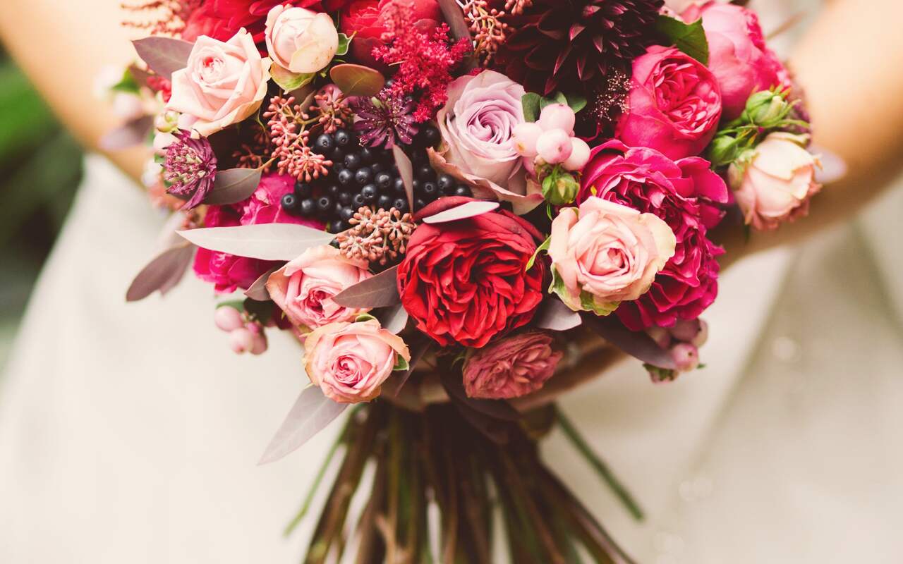A bridal bouquet of red and blush roses held against an ivory dress